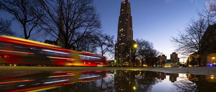 evening photo of Pitt campus, motion blur of bus moving in front of the Cathedral of Learning
